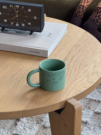 A Donna Wilson Tree Mug by Donna Wilson, in moss-green ceramic with a debossed tree design, sits on a wooden table near a clock and closed book as someone in patterned socks rests their feet nearby.