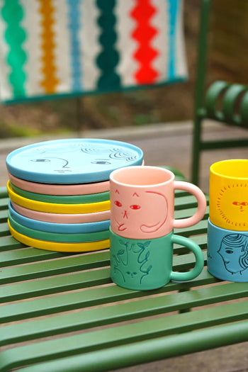 A Donna Wilson Tree Mug sits atop colorful ceramic plates on a green metal table. Behind them, a multicolored patterned cloth hangs near a matching green chair.