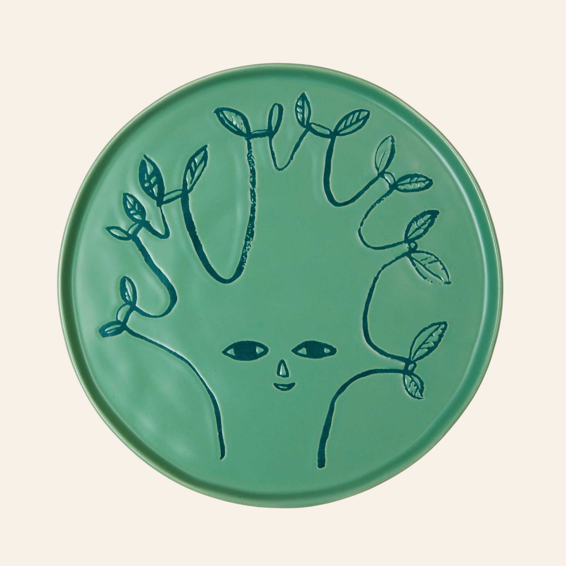 The Donna Wilson Tree Side Plate by Donna Wilson is a moss-green ceramic plate featuring a face with five branch-like arms adorned with small leaves, set against a plain beige background in a charming tree motif.