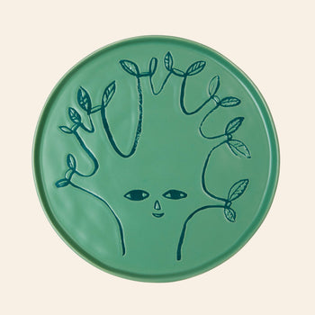 The Donna Wilson Tree Side Plate by Donna Wilson is a moss-green ceramic plate featuring a face with five branch-like arms adorned with small leaves, set against a plain beige background in a charming tree motif.