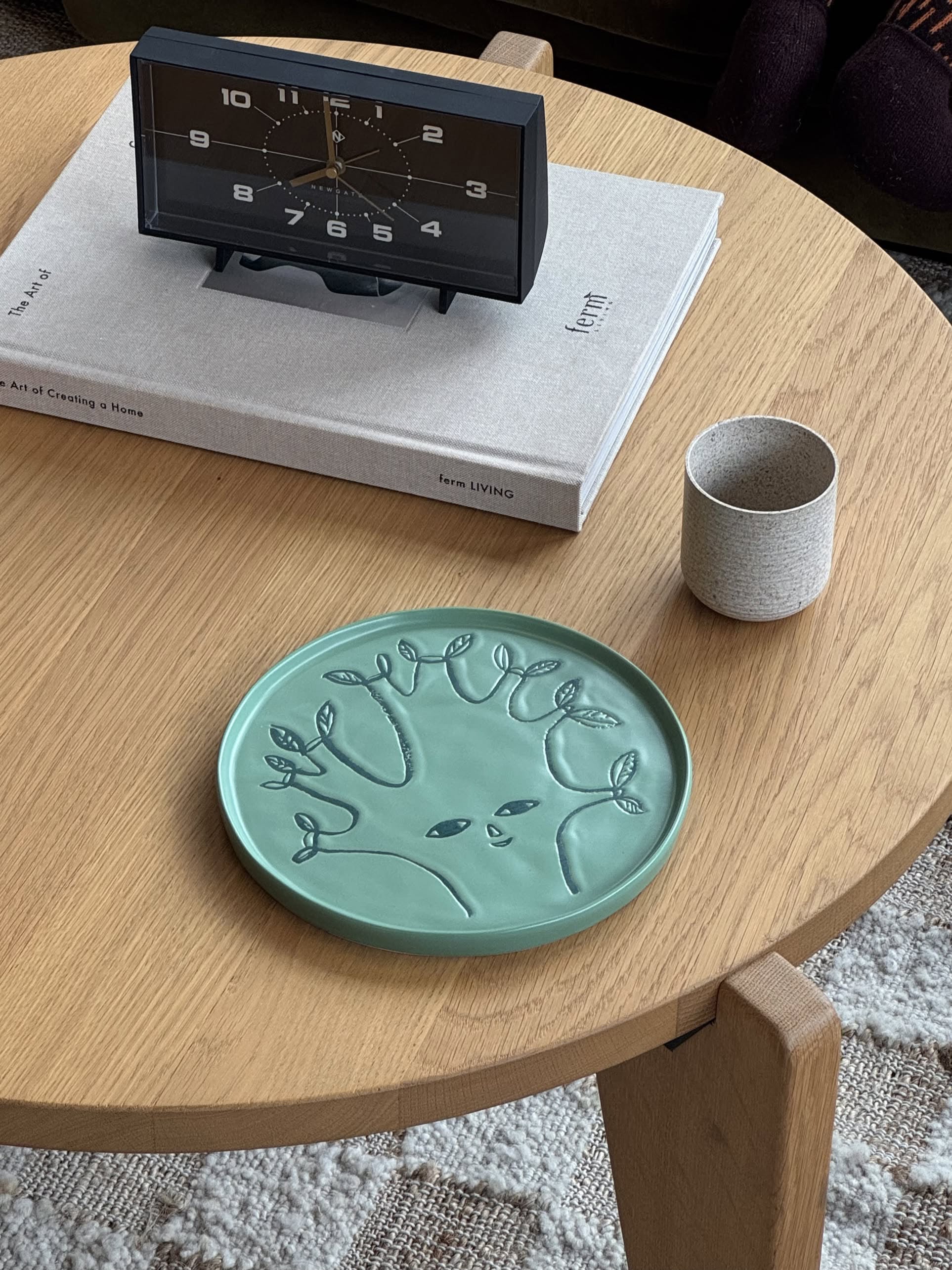 A round wooden table displays the Donna Wilson Tree Side Plate by Donna Wilson, a moss-green ceramic plate with a tree motif, alongside a small grey cup, a book topped with a black rectangular clock, and part of a patterned rug below.