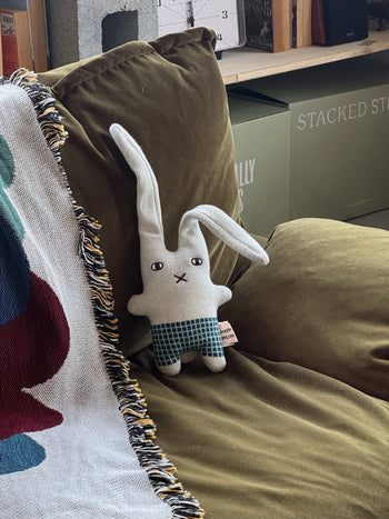 The Donna Wilson Wee One - Bonnie Bunny, a hand-embroidered stuffed bunny by Donna Wilson in blue checkered shorts, sits on a green couch beside a fringed patterned blanket. Shelves and boxes can be seen in the background.