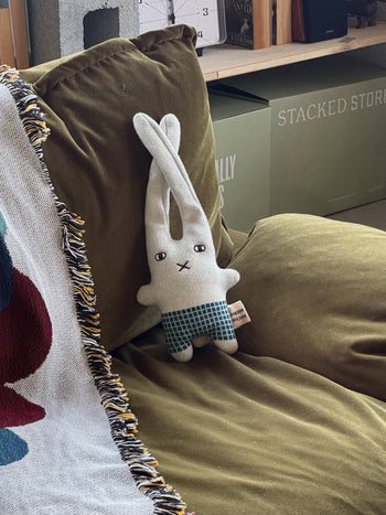 The Donna Wilson Wee One - Bonnie Bunny, a hand-embroidered organic cotton soft toy by Donna Wilson, sits on an olive green couch next to a fringed blanket, with books and storage boxes in the background.