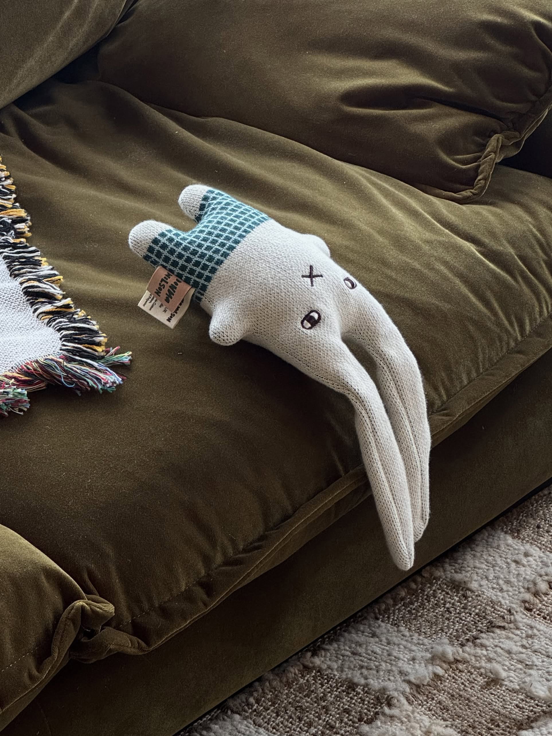 The Donna Wilson Wee One - Bonnie Bunny, a hand-embroidered organic cotton toy with long ears and a blue checkered shirt, lies on the edge of an olive green couch beside a fringed blanket.