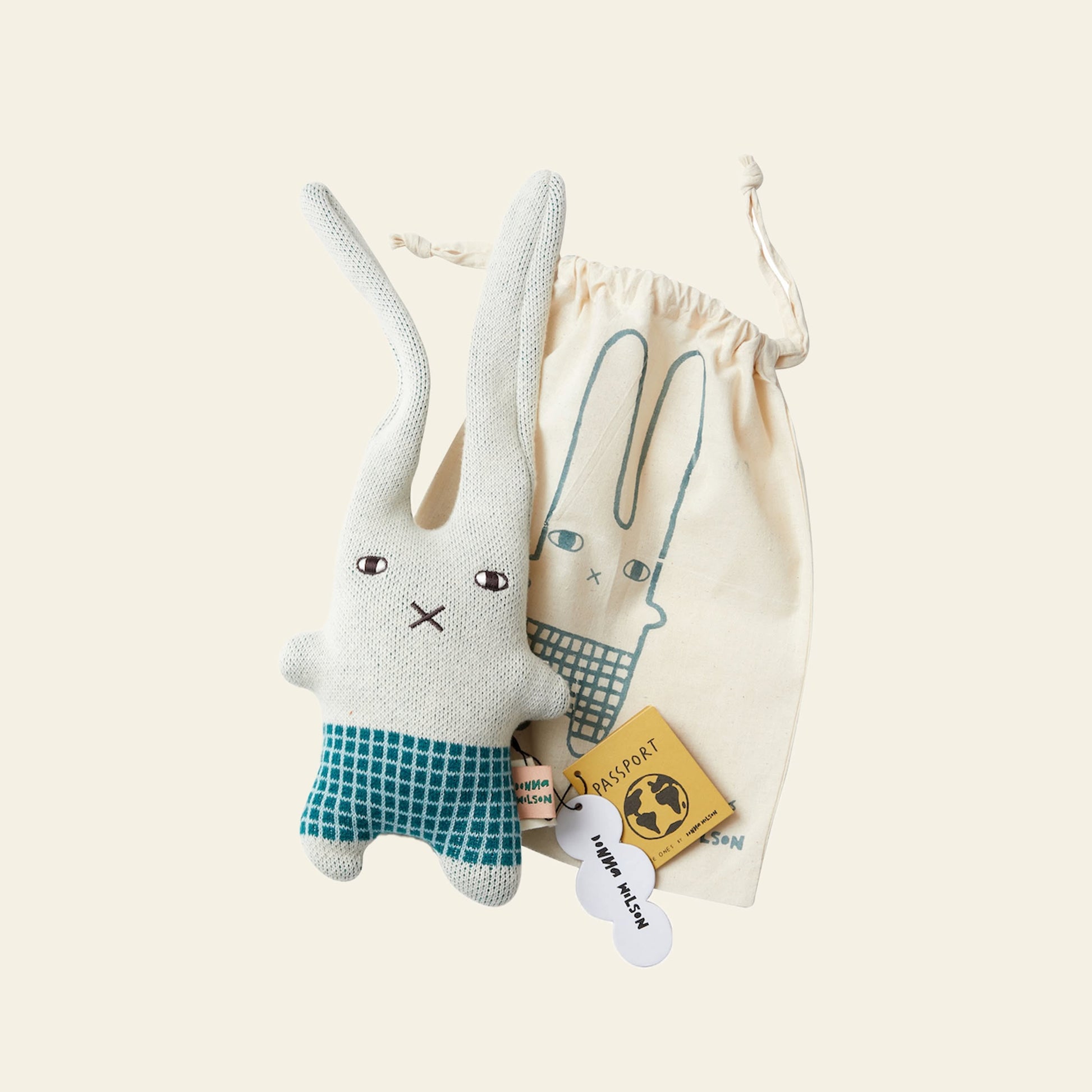 The Donna Wilson Wee One - Bonnie Bunny, a soft toy in green checkered shorts, rests by a matching bunny-illustrated drawstring bag, plus a yellow passport-shaped tag and a white bone-shaped tag.