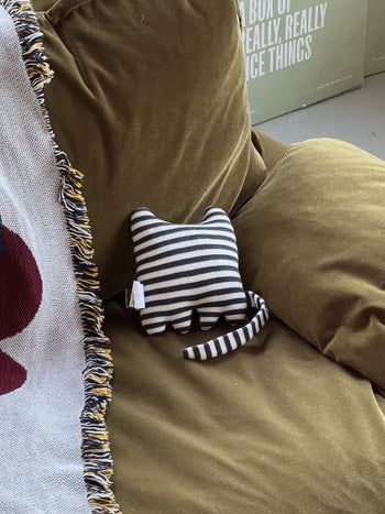The Donna Wilson Wee One - Mono Kitten, a hand-embroidered striped cat plush made from organic cotton yarn, rests on an olive green couch beside a fringed blanket.