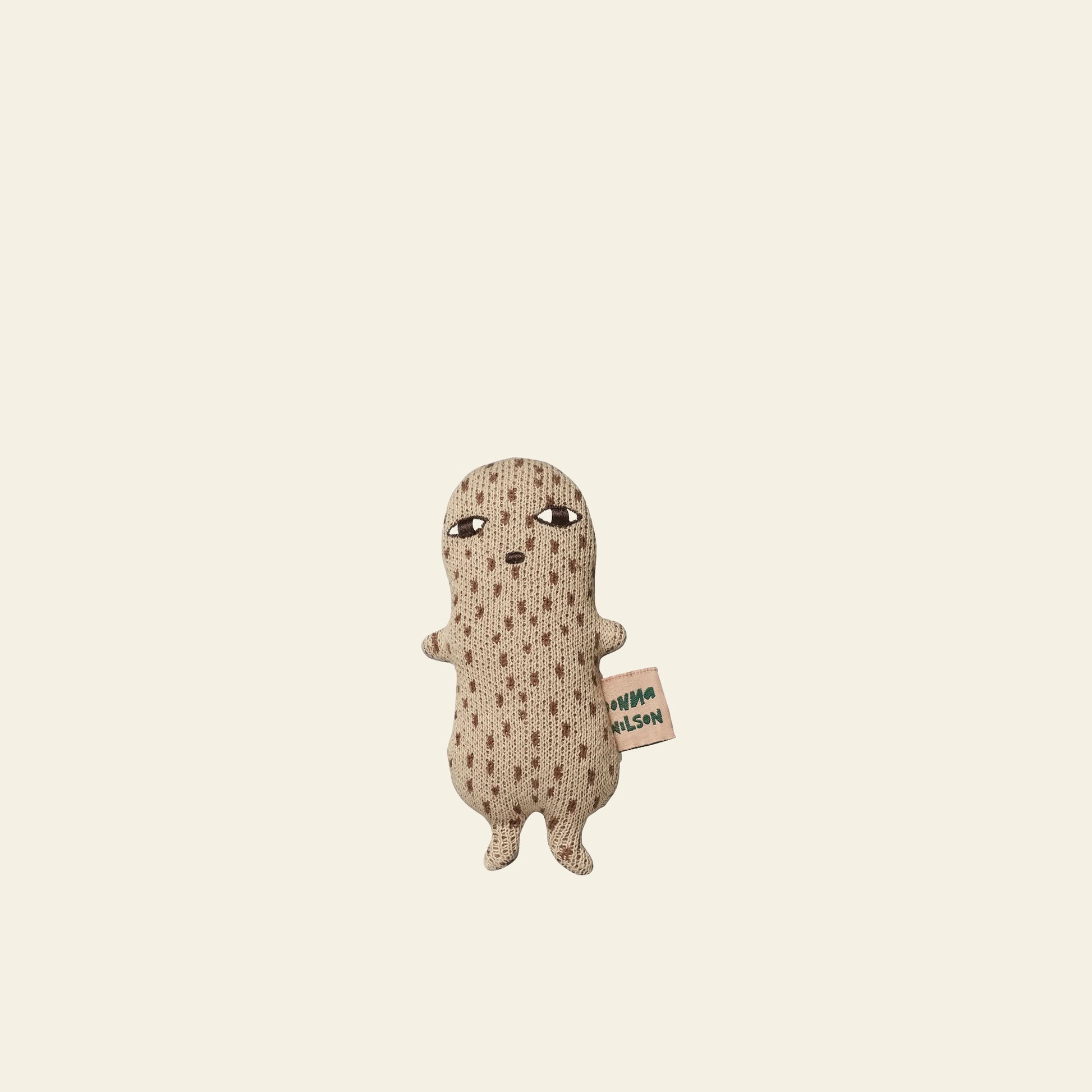 The Donna Wilson Wee One - Nutty by Donna Wilson is a small beige plush toy made from organic cotton, featuring dotted patterns, two arms, two legs, a neutral face, and a side tag. Stands upright against a plain background. Suitable from birth.