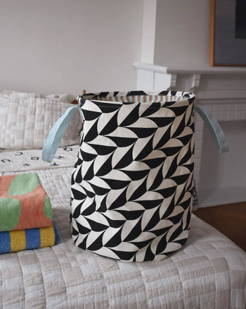 The Dusen Dusen Canvas Laundry Hamper by Dusen Dusen features a black and white scallop print, blue handles, and a durable PE-coated interior. It sits on a bed next to a folded towel, with a white fireplace and framed art in the background.