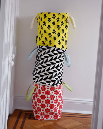 Three Dusen Dusen Canvas Laundry Hampers are stacked: the top has a yellow scallop print with black florals, the middle is black and white geometric, and the bottom is white with a red flower pattern.