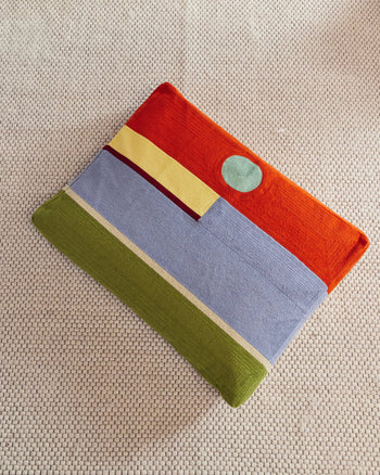 The Dusen Dusen Desert Dog Bed by Dusen Dusen features a colorful fabric laptop sleeve with abstract shapes in orange, green, yellow, light blue, and a teal circle, resting on a textured beige surface beside the cozy dog bed.