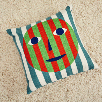 The Dusen Dusen Everybody Pillow Cover by Dusen Dusen sits on a beige carpet, showcasing a bold multi-coloured embroidered face with striped red and green lines, blue eyes and mouth, on a white and blue striped background.