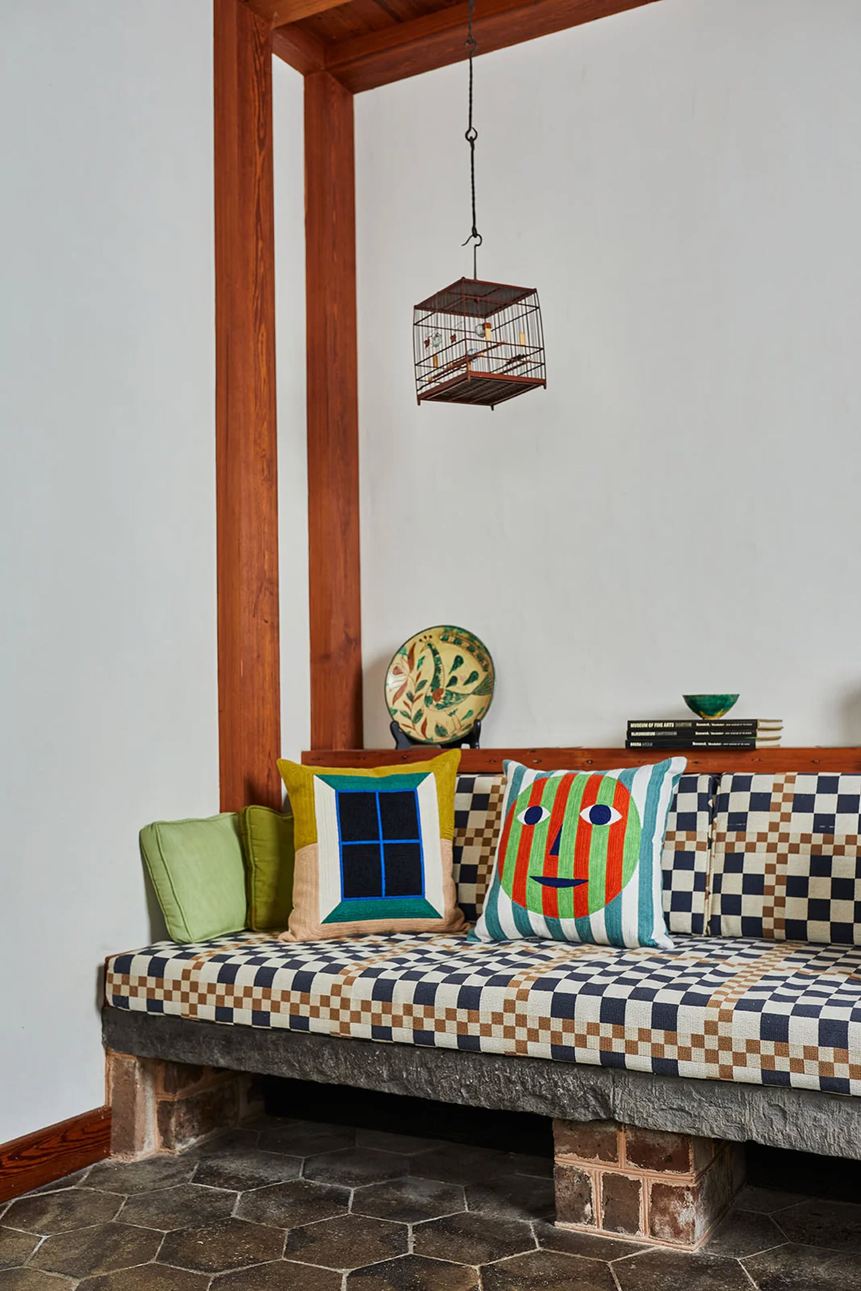 A checkered mosaic bench with colorful Dusen Dusen Everybody Pillow Covers rests against a white wall. Above, a small birdcage hangs, while a shelf behind displays stacked books and a decorative plate.