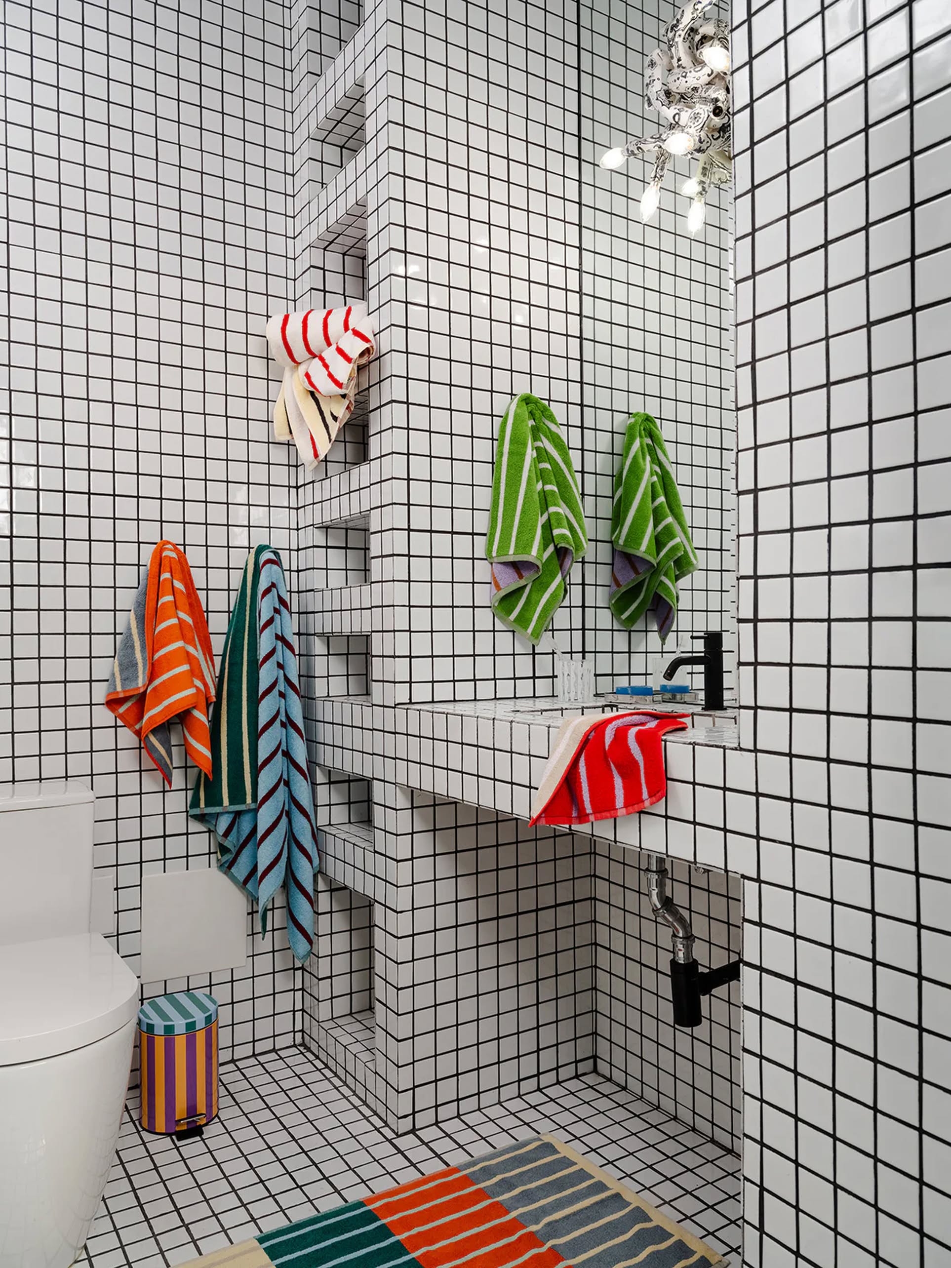 A modern bathroom features white square tiles with black grout. Dusen Dusen House Stripe Glass Hand Towels bring vibrant color, and a matching striped rug plus a multicolored waste bin complete the lively look.