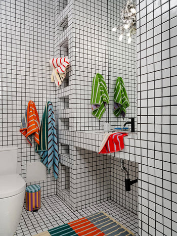 A modern bathroom with white square tiles is brightened by pops of color from a Dusen Dusen House Stripe Stone Hand Towel and matching rug, while a soft towel and striped waste bin complete the lively look.