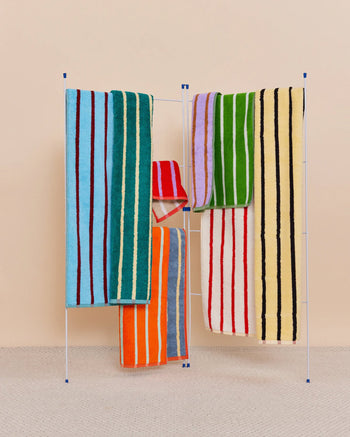 A drying rack displays five Dusen Dusen House Stripe Stucco Bath Towels, featuring bold stripes in blue, green, yellow, orange, red, and purple on a beige background. A light grey textured floor complements the colorful display.