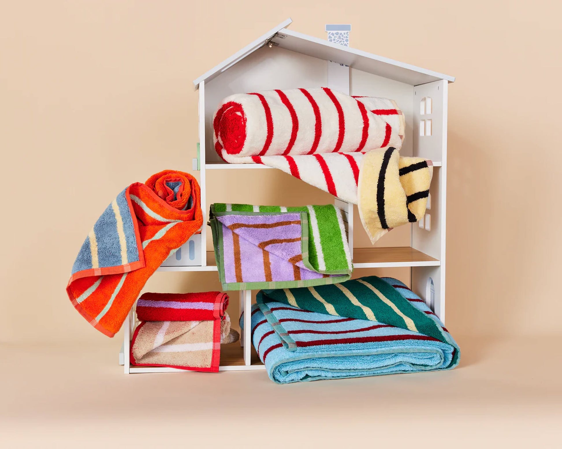 The Dusen Dusen House Stripe Stucco Bath Towels by Dusen Dusen, in bold striped colors, are rolled or folded on a dollhouse’s floors and roof set against a beige background.