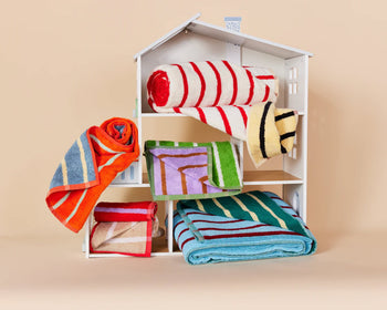 A white dollhouse showcases five Dusen Dusen House Stripe Tile Bath Towels in red, orange, blue, green, and yellow—draped or rolled throughout different levels—set against a beige background. Brand: Dusen Dusen.