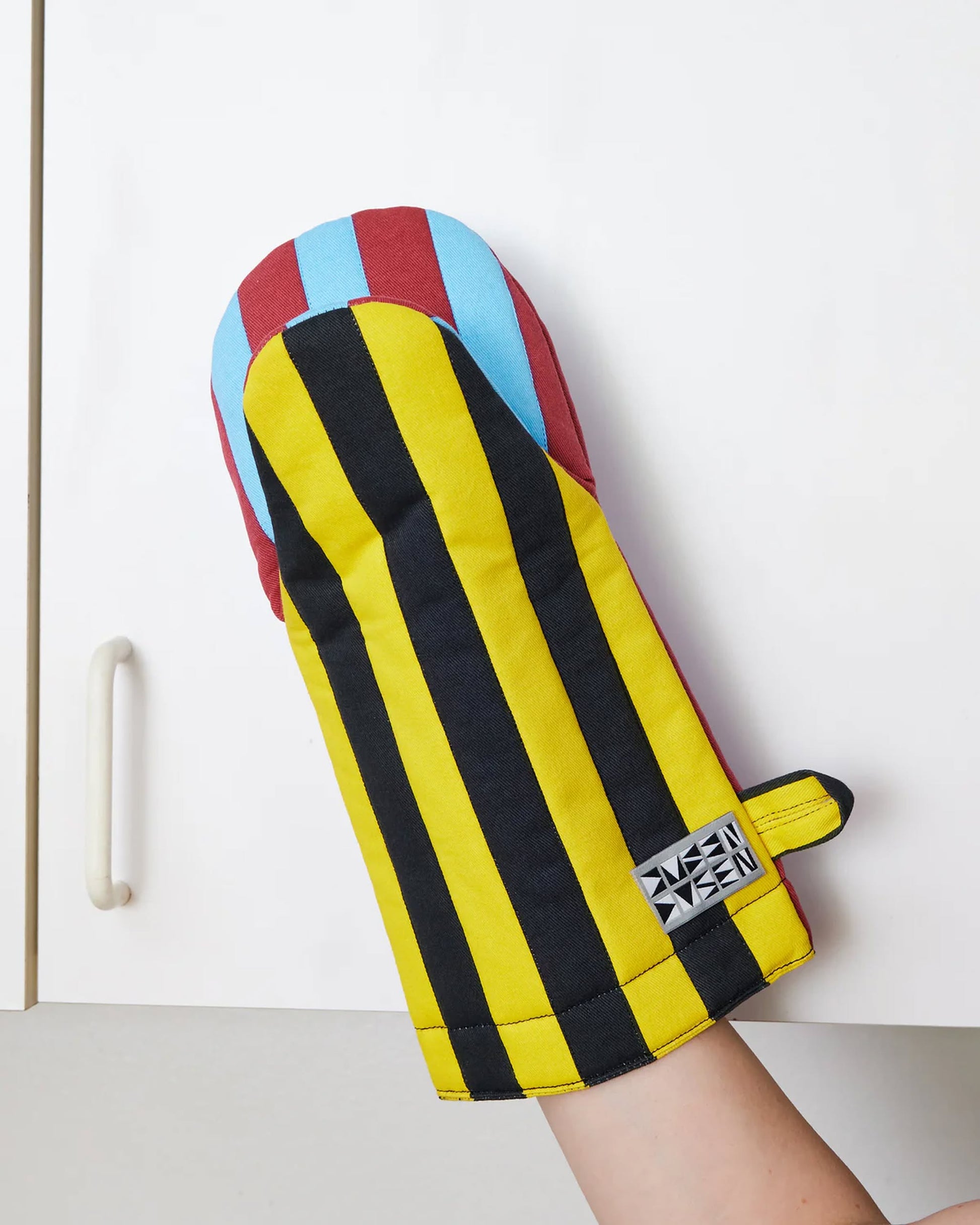 A hand holds up the Dusen Dusen Striped Oven Mitt by Dusen Dusen, featuring colorful stripes—including vertical yellow, black, red, and blue on the thumb—in front of a white cabinet.