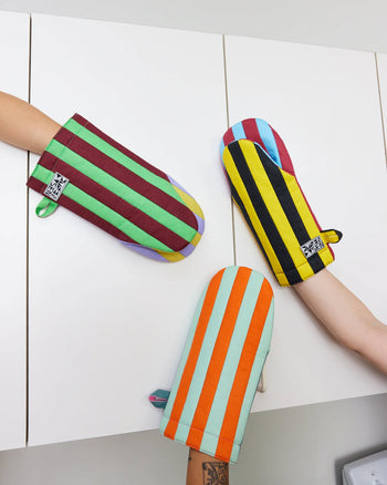 Three arms wearing Dusen Dusen Striped Oven Mitts, a vibrant kitchen essential from Dusen Dusen, reach toward each other in front of white cabinets.