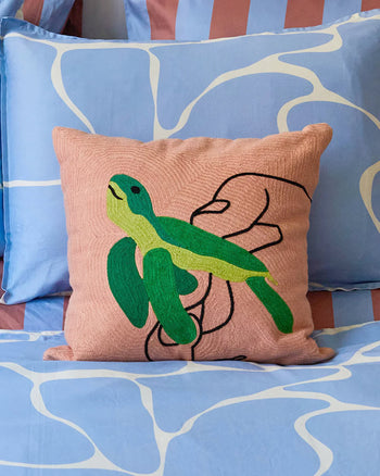 The Dusen Dusen Turtle Pillow Cover, in pink with a green turtle embroidery, is shown held by hand and displayed on blue and white abstract sheets—a playful accent by Dusen Dusen for marine-inspired decor.