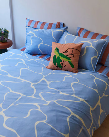 A neatly made bed with blue and white bedding features the Dusen Dusen Turtle Pillow Cover, adding a playful marine touch. Matching pillows and a small plant on the bedside table complete the cozy decor.