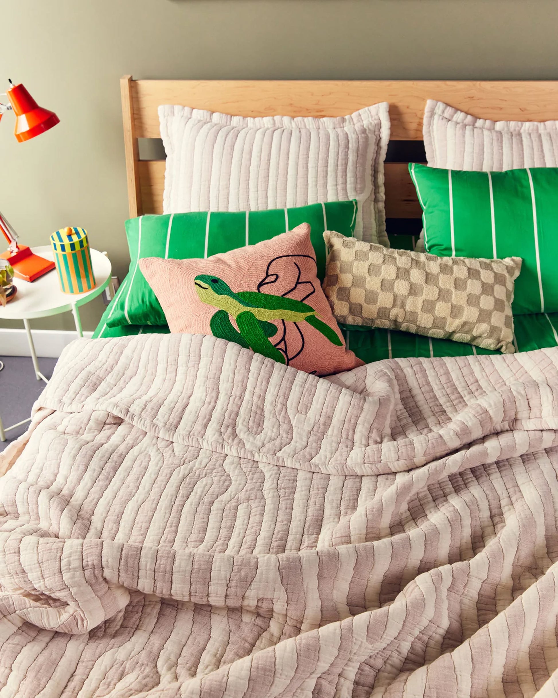 A neatly made bed features green-striped pillows, the Dusen Dusen Turtle Pillow Cover by Dusen Dusen with a geometric pattern, and a pink pillow with a green bird. Light pink textured bedding is paired with marine life decor on the nightstand.
