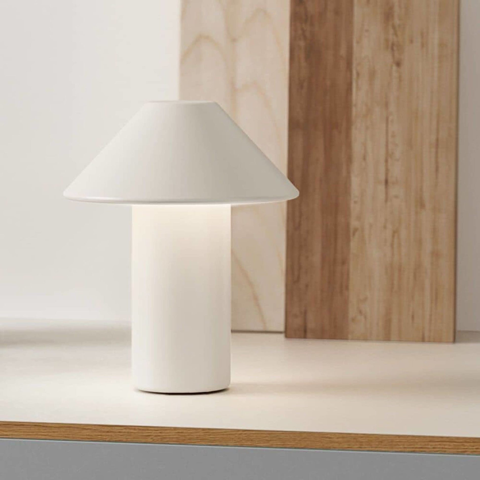 The EDGAR ROY Lamp by EDGAR, a modern white table lamp with a cylindrical base and wide conical shade, sits on a light-toned surface against wooden paneling in the background.