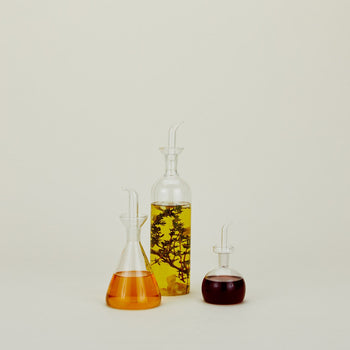 Three glass bottles in various shapes and sizes, including the Hawkins New York Essential Kitchen Bottle, Medium, are filled with colored liquids—clear yellow oil with herbs, amber, and dark red—and set against a plain light background.