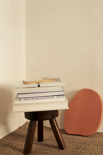 A compact FRAMA AML Stool in solid oiled pine holds books and magazines in a minimalist room with beige walls and a woven rug. Nearby, a flat, reddish-brown decorative piece rests against the wall.