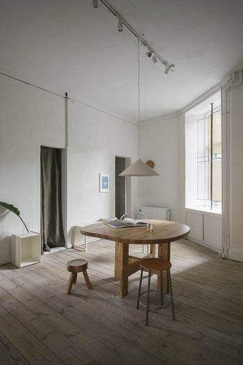 Minimalist room with a round wooden table, a solid oiled pine FRAMA AML Stool by FRAMA beside the bed, another stool, pendant lamp, plant in the corner, light wood floors, curtained doorway, and a tall window filling the space with natural light.