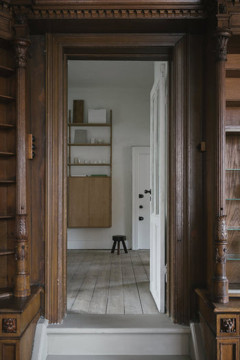 A view through a carved wooden doorway into a minimalist room with light wood floors, white wall, shelves, cabinet, and the FRAMA AML Stool by FRAMA—crafted from solid oiled pine—placed near a white door with black handles.