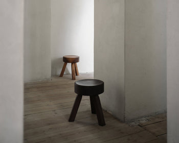 Two FRAMA AML Stools by FRAMA, one in dark and one in light solid oiled pine, stand in separate corners of a minimalist room. Their versatile design suits use as bedside stools or accent pieces, enhancing the space’s calm and simple vibe.