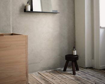 A minimalist bathroom with light wooden floors features a solid oiled pine cabinet and the FRAMA AML Stool by FRAMA as a side table holding a soap dispenser. A wall-mounted shelf with mirror enhances the versatile design, illuminated by window light on the right.