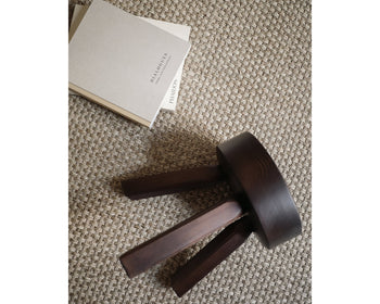 A compact, dark solid oiled pine FRAMA AML Stool by FRAMA rests on its side atop a textured beige carpet beside two closed, neutral-toned books, one titled DIALOGUES.