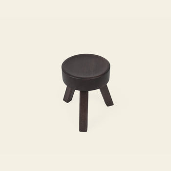 The FRAMA AML Stool by FRAMA is a small, round, dark wooden bedside stool with three angled legs, crafted from solid oiled pine and shown against a plain off-white background.