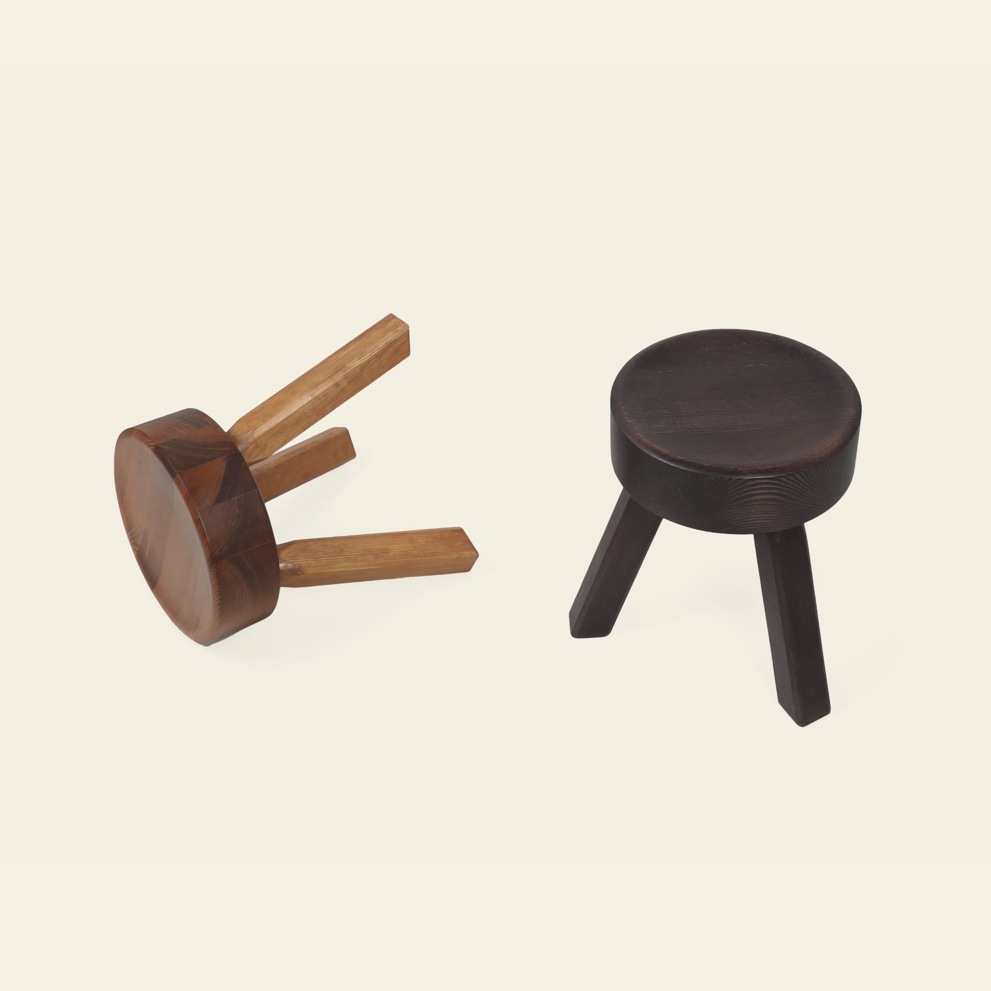 Two FRAMA AML Stools by FRAMA are displayed on a light background; one stands upright with dark wood legs, while the other, crafted from solid oiled pine, is tipped over to showcase their versatile three-legged design.