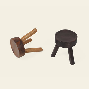 Two FRAMA AML Stools by FRAMA are displayed on a light background; one stands upright with dark wood legs, while the other, crafted from solid oiled pine, is tipped over to showcase their versatile three-legged design.