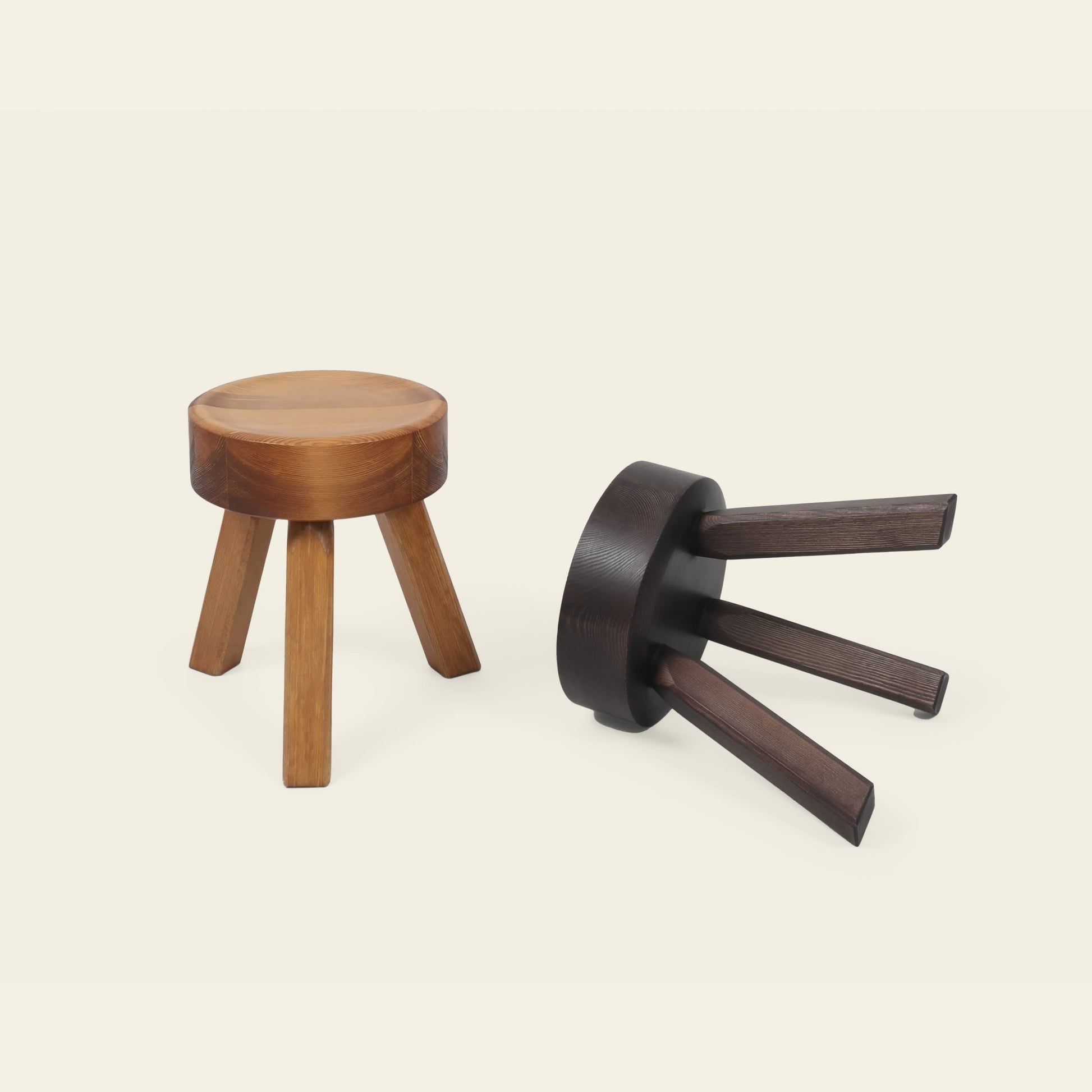 Two FRAMA AML Stools by FRAMA—one upright in natural oiled pine, one tipped over in dark wood—are displayed on a light background, highlighting their versatile design as bedside stools or extra seating.