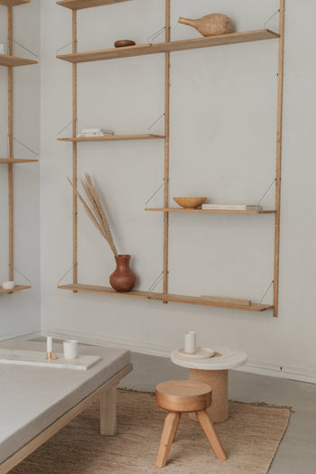A minimalist living space features wooden wall shelves with pottery, a brown vase, bowls, books, and candles. The versatile FRAMA AML Stool by FRAMA serves as a bedside table alongside a round table and a light floor rug.