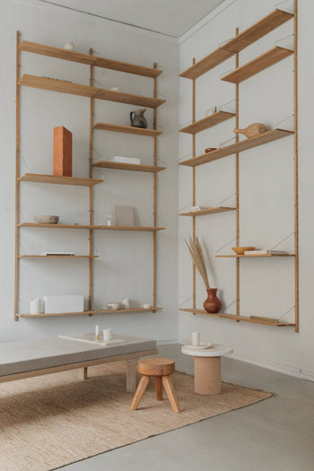 Minimalist living room with light wood wall shelves, a low cushioned bench, and a round side table holding a vase of dried grass. The versatile FRAMA AML Stool stands out beside a jute rug on the concrete floor.