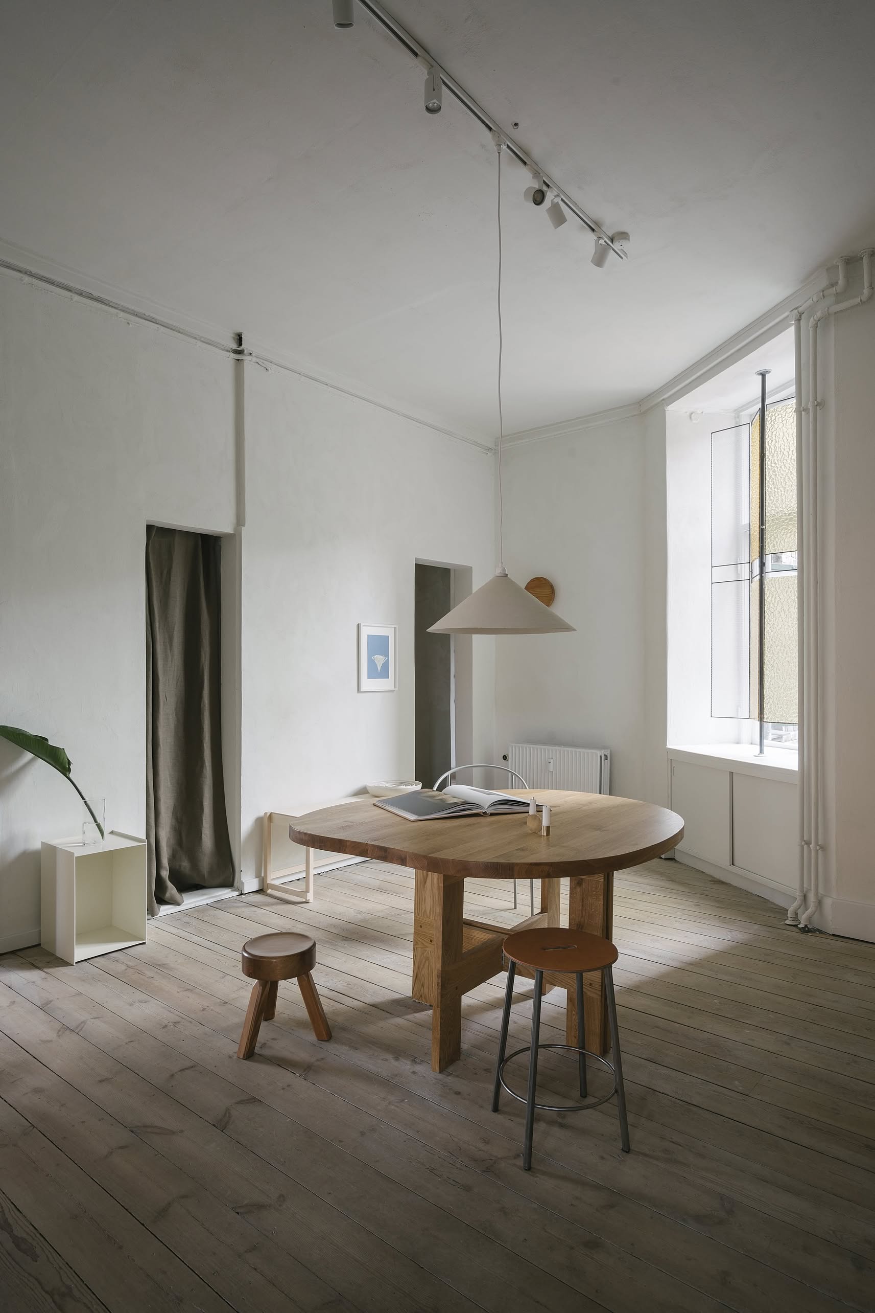 Minimalist dining room with a round wooden table, two FRAMA AML Stools in solid oiled pine, a pendant lamp, light wood floors, a leafy plant, and soft natural light. The FRAMA stools versatile design keeps the space simple and modern.