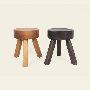 Two FRAMA AML Stools by FRAMA are shown side by side: one in light brown oiled pine, the other in dark brown. Both have round, thick seats and angled legs—versatile for use as a bedside stool or extra seating.