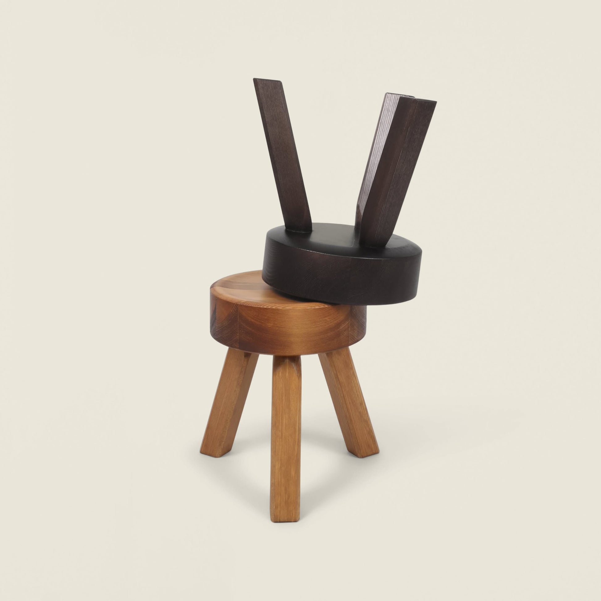 Two FRAMA AML Stools by FRAMA—solid oiled pine, one light brown and three-legged, the other dark brown and tilted—are abstractly stacked to highlight their versatile design, perfect as bedside stools.