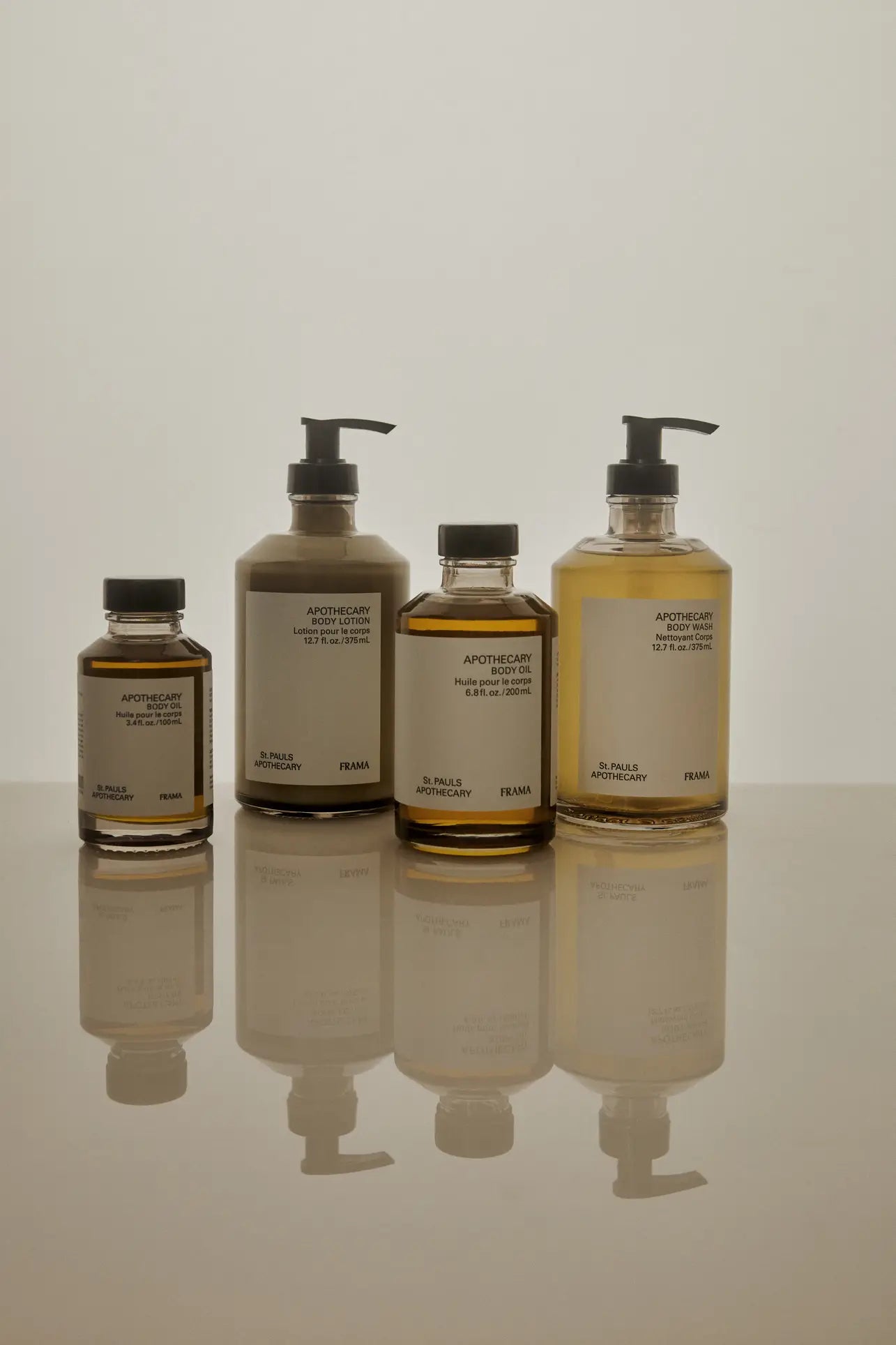 Four minimalist FRAMA Body Wash bottles (375ml) with pump and screw caps are lined up on a reflective surface against a beige background. Each is labeled FRAMA and contains soothing or fragrant cleansing gel.