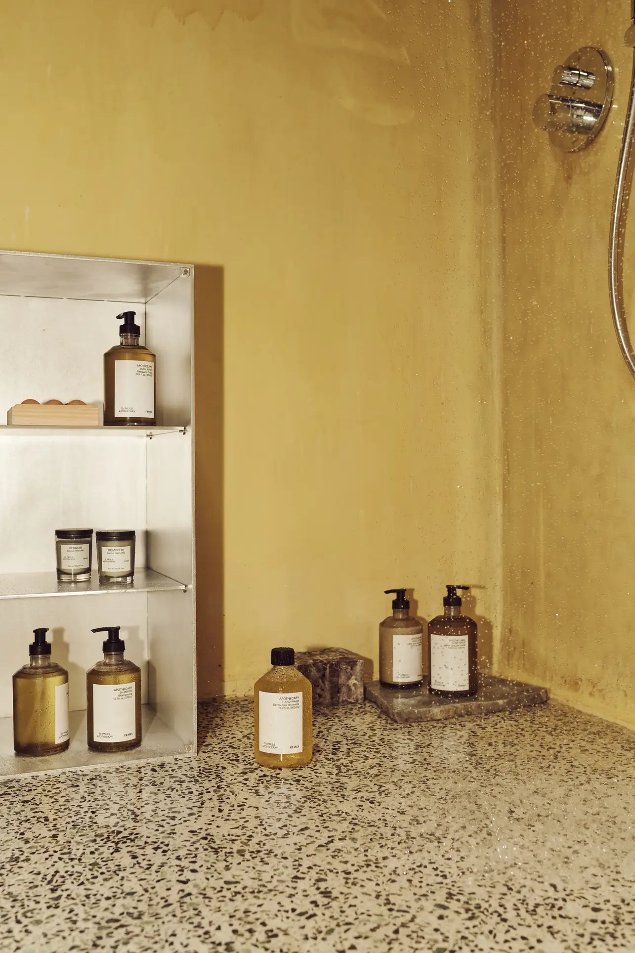 A minimalist shower with yellow walls, terrazzo floor, and a metal shelf holding bottles and jars—including FRAMA Body Wash, 375ml—plus more bath products arranged near the shower tap.
