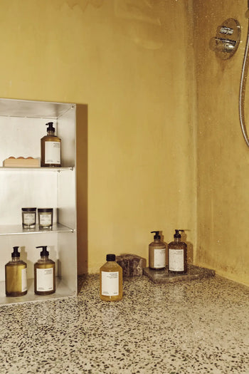 A minimalist shower with yellow walls, terrazzo floor, and a metal shelf holding bottles and jars—including FRAMA Body Wash, 375ml—plus more bath products arranged near the shower tap.