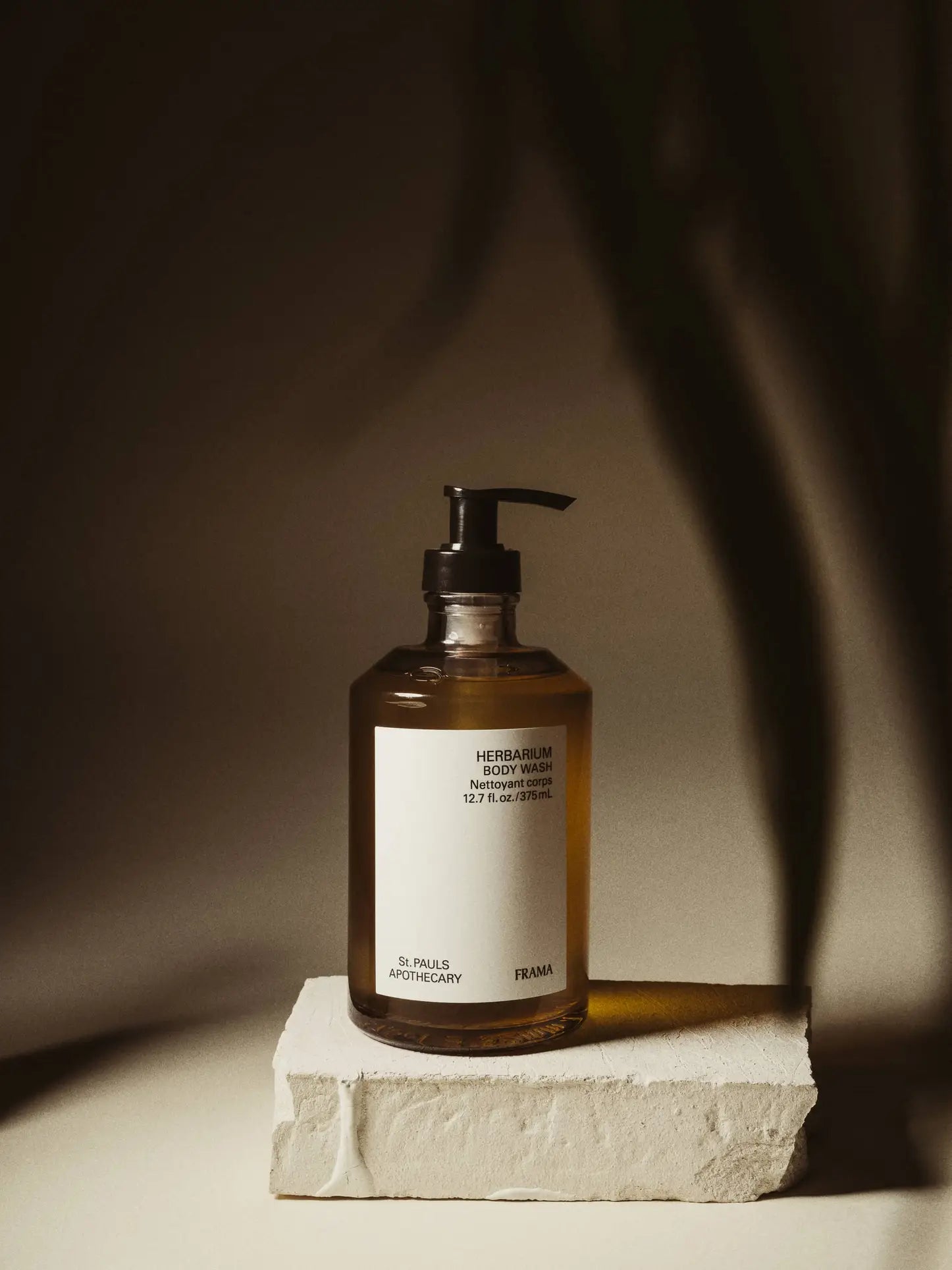 A brown pump bottle labeled FRAMA Body Wash, 375ml by FRAMA sits on a rectangular white stone against a beige background, its gentle shadows emphasizing the calming vibe of this aromatic gel in a warm, minimalist setting.