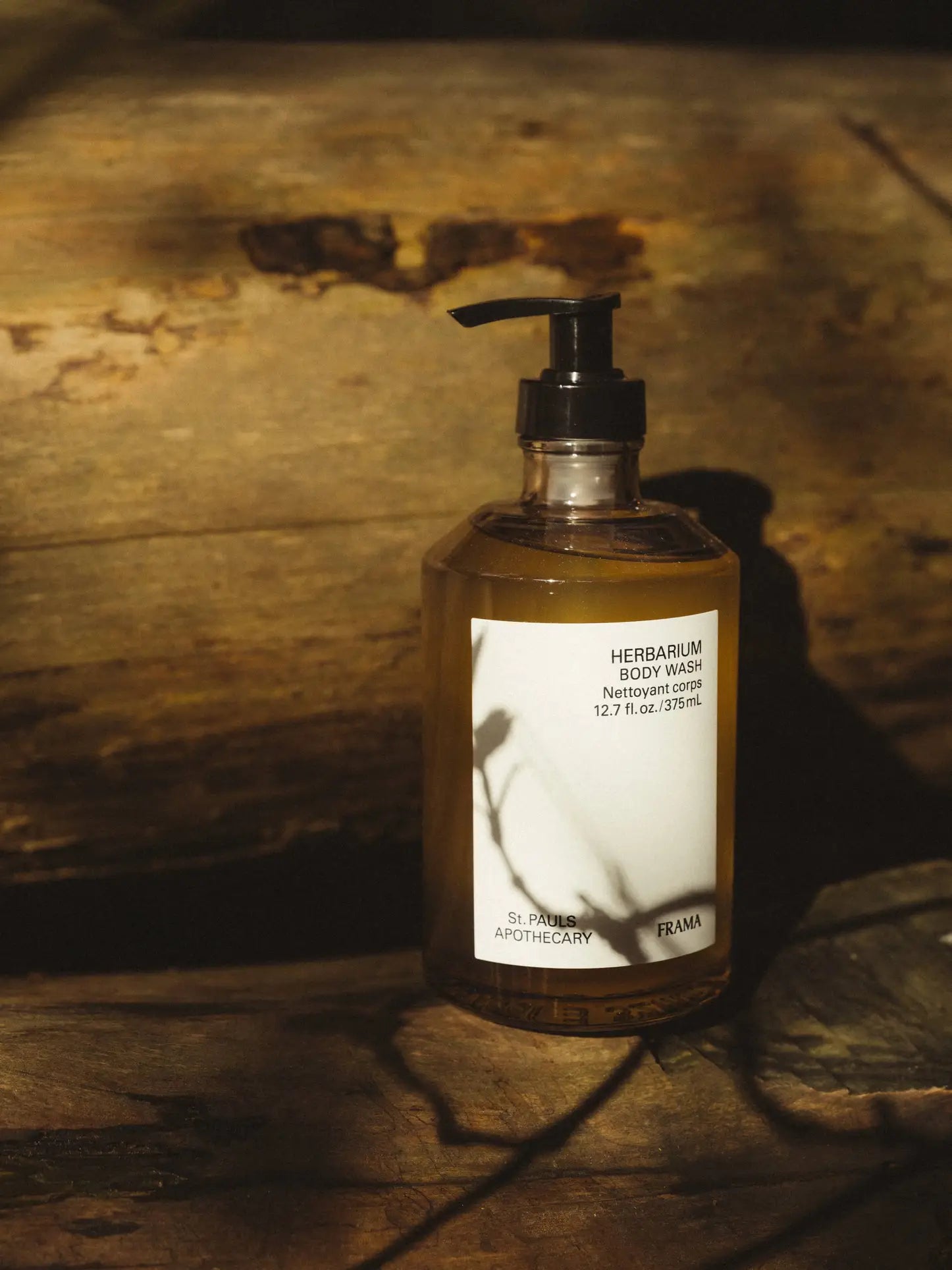 A bottle of FRAMA Body Wash, 375ml, with a pump top sits on a wooden surface in warm light. The label displays “FRAMA Body Wash 12.7 fl. oz./375 ml, St. Paul’s Apothecary.”.