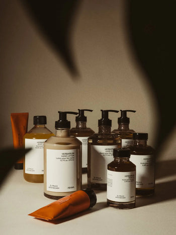 A selection of FRAMA personal care products, featuring the FRAMA Body Wash (375ml) in brown and orange bottles with white labels, arranged on a beige surface with shadows and dark shapes framing the scene.