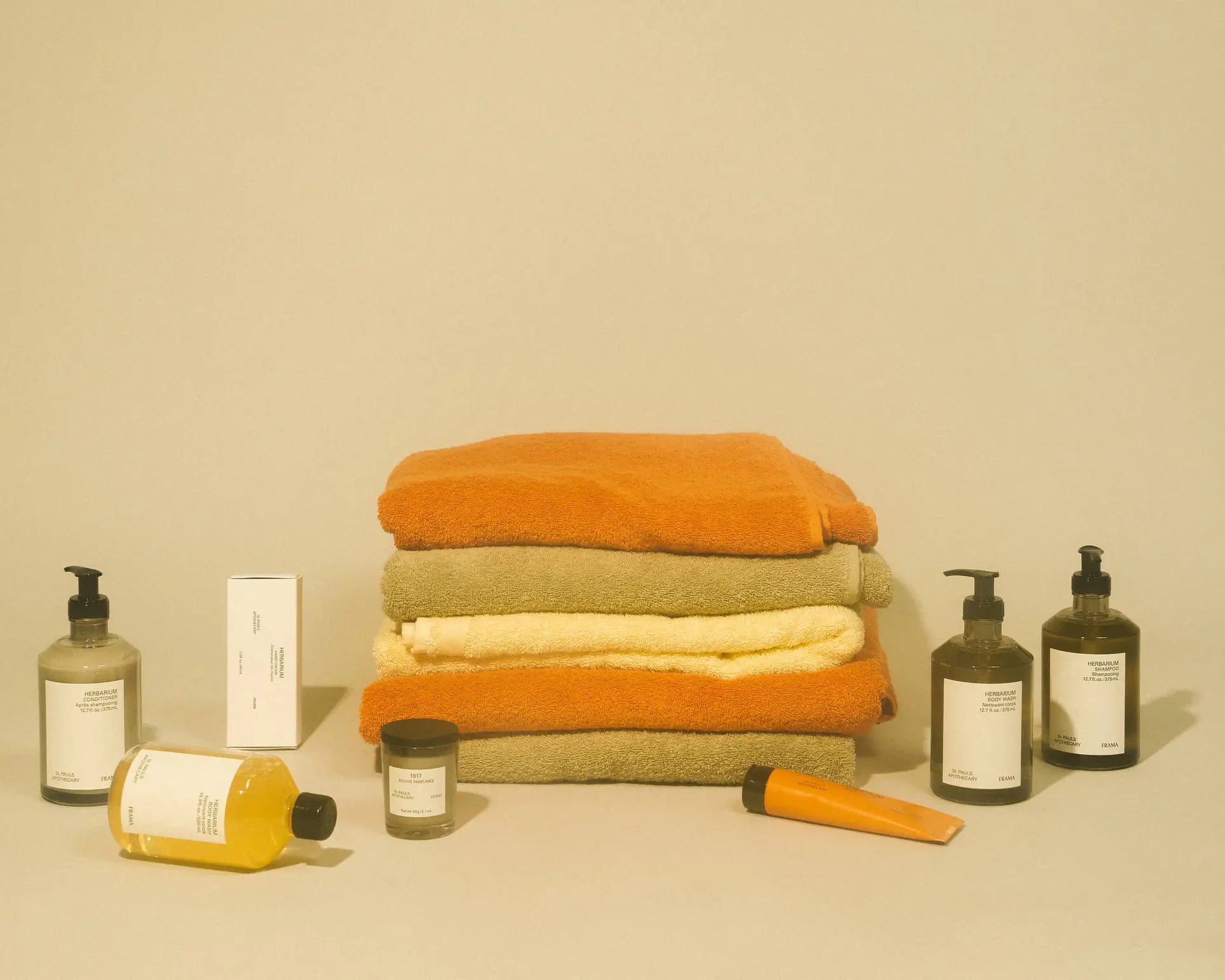 Three folded towels in orange, green, and yellow sit center stage on a beige background, surrounded by skincare bottles—including FRAMA Body Wash, 375ml—along with various jars and tubes featuring minimal labels.